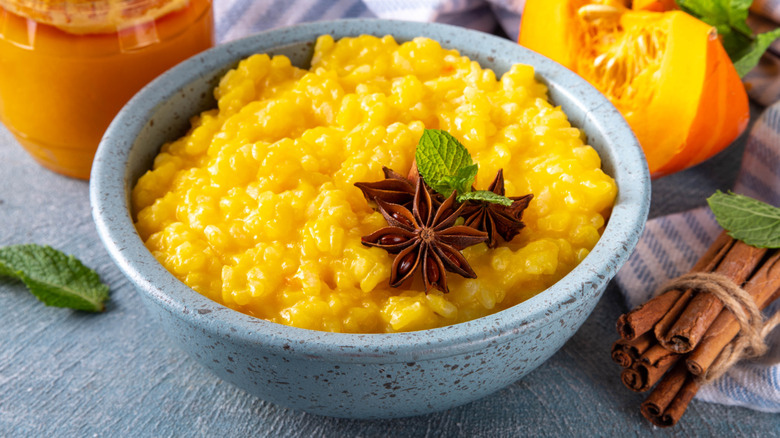 a stoneware bowl of pumpkin rice pudding garnished with mint and star anise, with cinnamon sticks, mint leaves, and a wedge of pumpkin beside the bowl