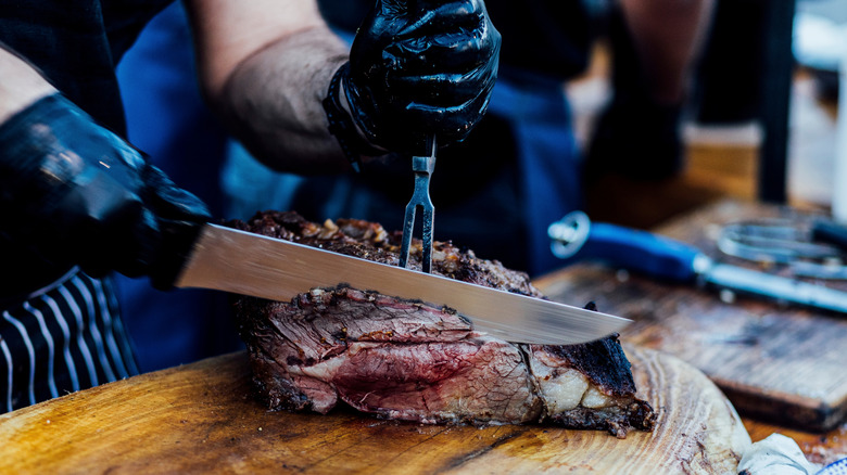 A worker with black nitrile gloved hands slices a large brisket