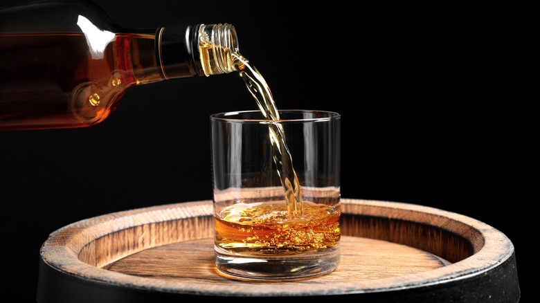A bottle pouring whiskey into a glass sitting on top of a wooden barrel