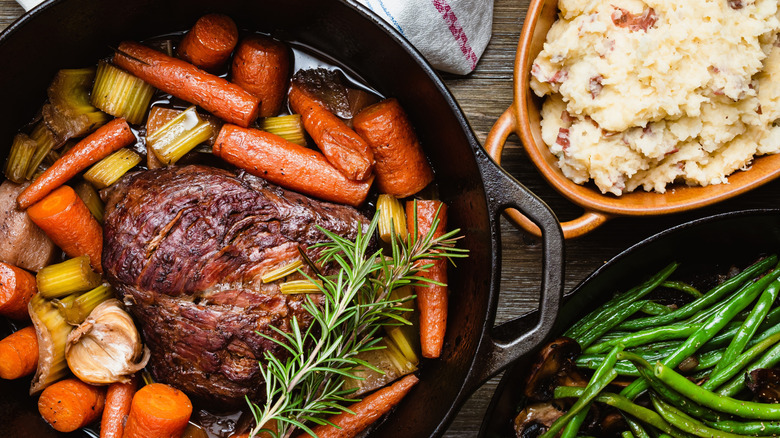 pot roast with carrots and celery in a cast iron pot beside a ceramic dish of mashed potatoes