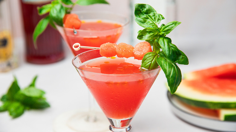 Two watermelon martini glasses with bottles and slices in background.