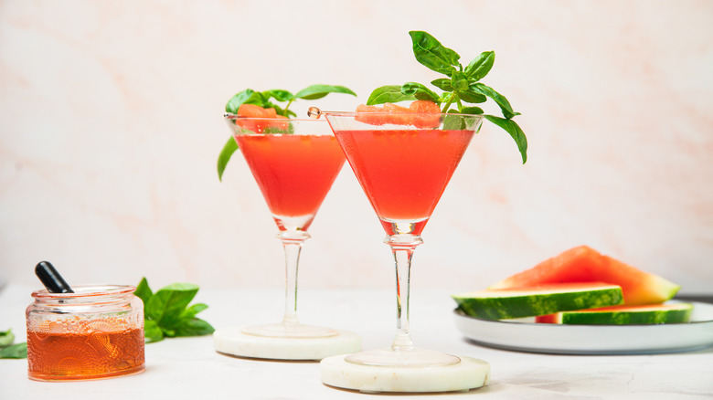 Two watermelon martini glasses with oil and slices in background.