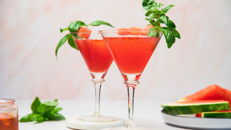 Two watermelon martini glasses with oil and slices in background.