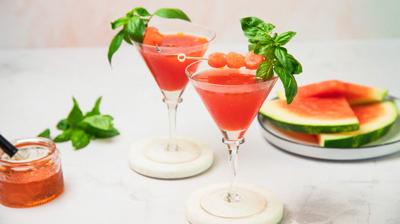 Two watermelon martini glasses with oil and slices in background.