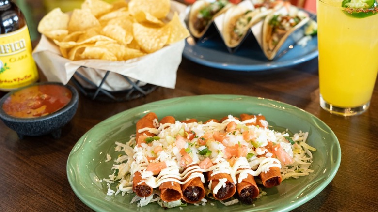 Platter of taquitos with chips, salsa, tacos, and cocktail in background