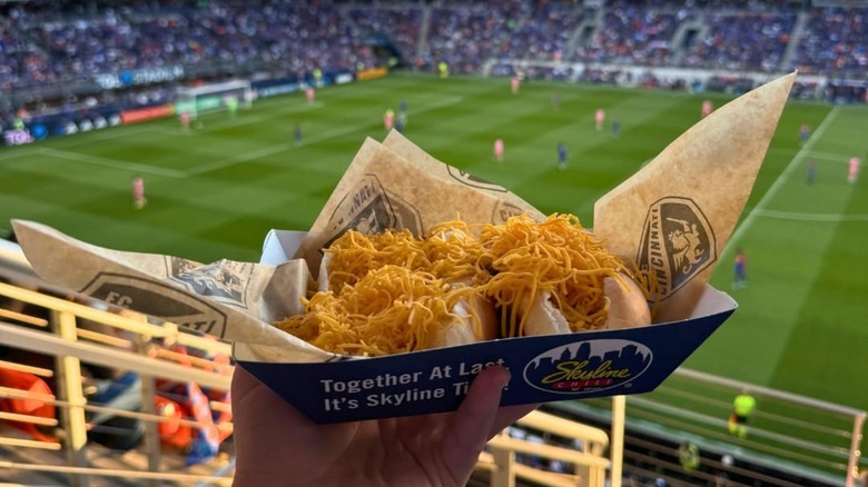 Hand holding two chili cheese hot dogs in Skyline Chili tray with soccer field blurred in background