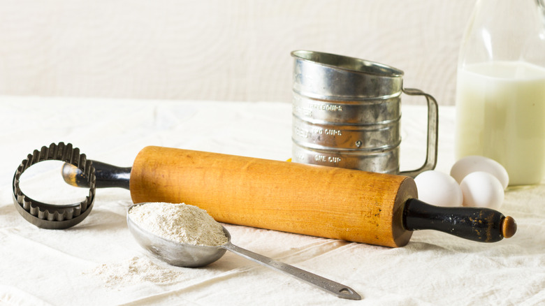 An old metal flour sifter beside a rolling pin, a measuring spoon with flour. eggs, a bottle of milk, and cookie cutters