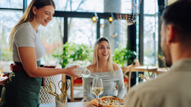 A smiling server pouring a glass of white wine for a happy couple at a bright and airy restaurant