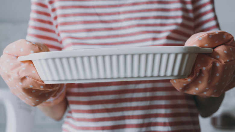 A close-up of a woman wearing oven mitts and holding out a white ceramic casserole dish