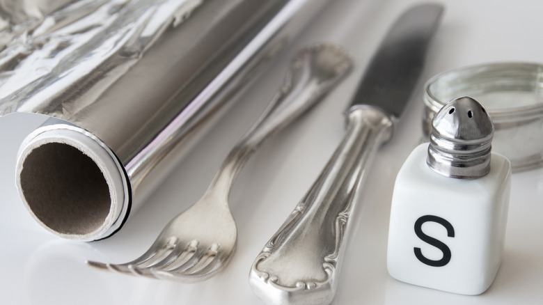 A roll of aluminum foil, a silver butter knife and fork, and a shaker of salt on a white countertop