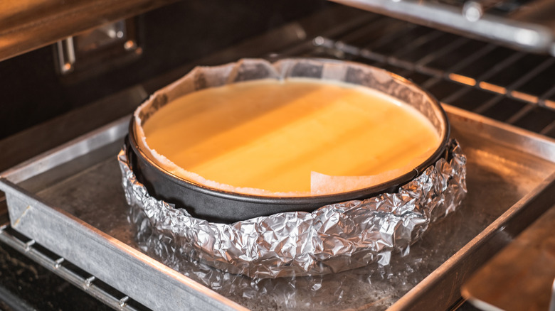A cheesecake baking in a springform pan that's been wrapped in aluminum foil and is resting in a water bath in the oven