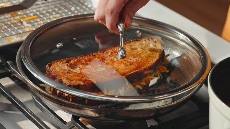 A crispy sandwich cooking in a saute pan with someone holding the lid on top