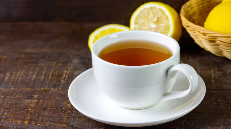 A cup of tea sits on a saucer, with a wicker basket of lemons and several sliced lemons in the background