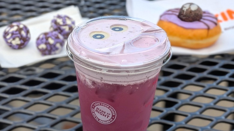 A Dunkin' refresher with candy eyeballs on top on a picnic table and a donut and Munchkins in the background
