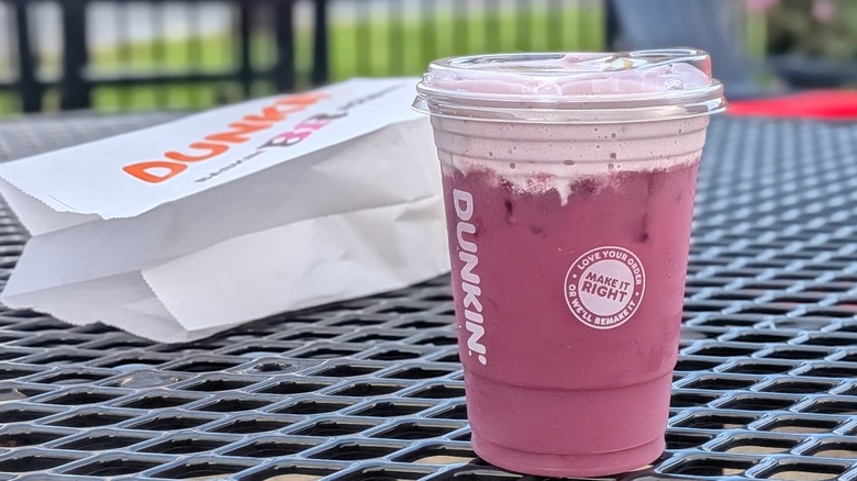 A purple refresher from Dunkin' with cold foam on an outdoor picnic table in front of a white Dunkin' branded pastry bag