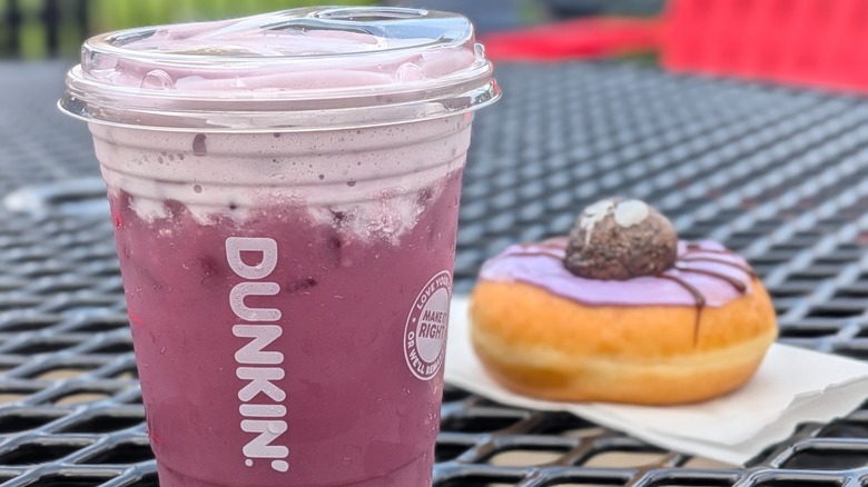 A close-up of a purple Dunkin' refresher with cold foam on a picnic table and a spider donut in the background