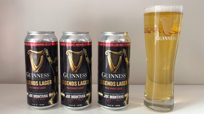 Three cans of Guinness Legends Lager next to tall Guinness pint glass on white table