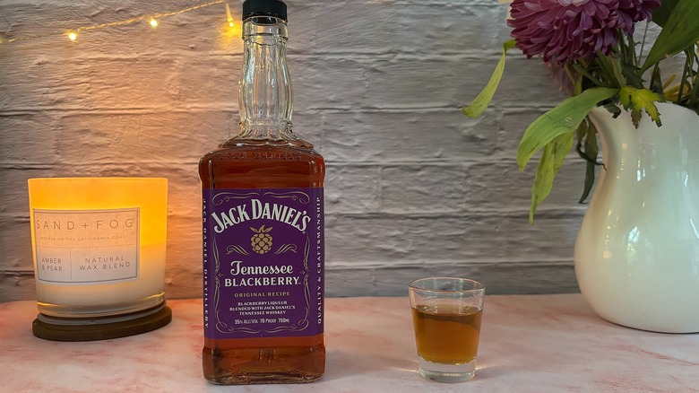 Shot glass of Jack Daniel's Tennessee Blackberry whiskey next to bottle of whiskey, candle and flowers in background