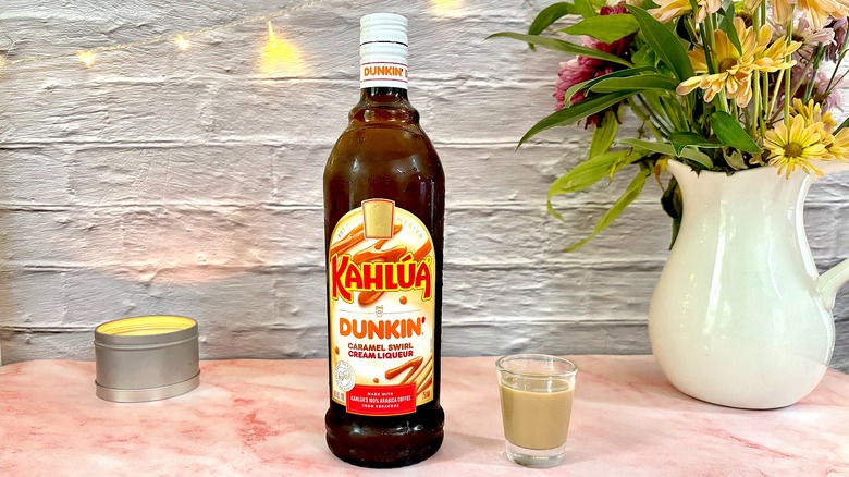 Shot glass of Kahlúa Dunkin Caramel Swirl Cream Liqueur next to bottle, with candle and flowers in background