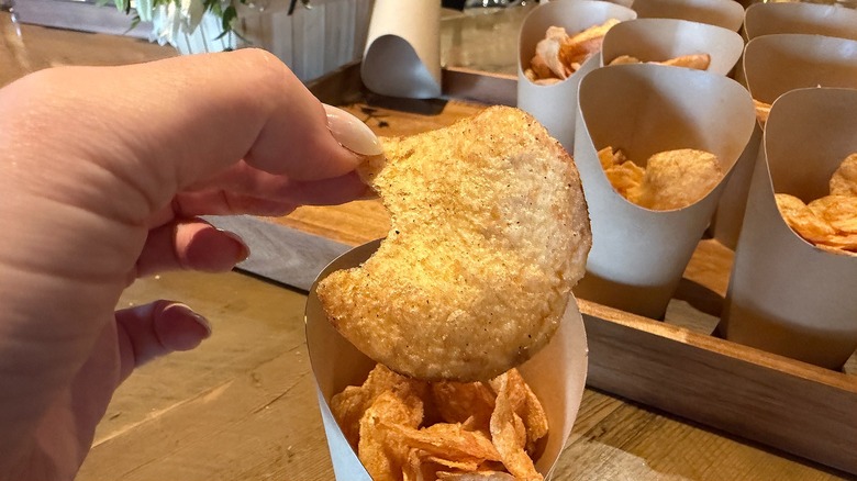 Hand holding Lay's chip above several cups of Lay's chips on wooden table