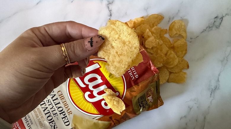 Hand holding Lay's Cheddar Scalloped Style Potato chip above bag of chips with chips spilling out onto marble surface