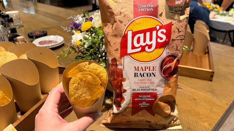 Bag of Lay's Roasted Maple Bacon potato chips next to cup of chips on wooden table, more cups of chips beside