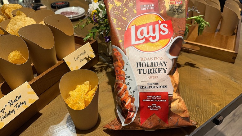 Bag of Lay's Roasted Holiday Turkey potato chips next to cup of chips on wooden table, more cups of chips beside