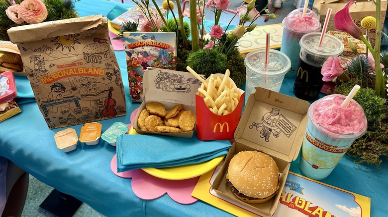 A McDonaldland Meal on a table set with flowers, including a bag, sauce packets, 10-piece Chicken McNuggets, a souvenir character tin, fries, a quarter pounder with cheese, and a Mt. McDonaldland shake set next to a yellow plate with a blue napkin.