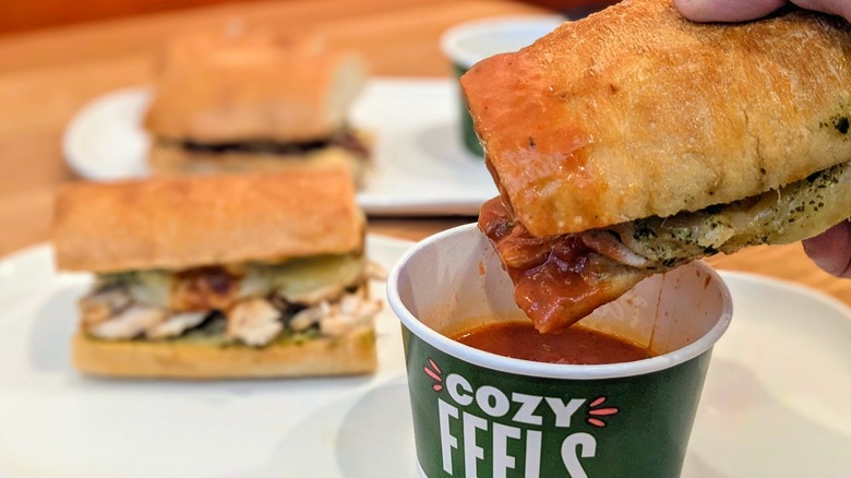 A close-up of half a sandwich being dunked into tomato soup with sandwiches blurred in the background