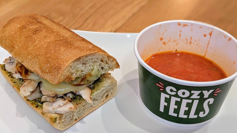 Half a chicken and pesto sandwich next to a cup of tomato soup on a white plate