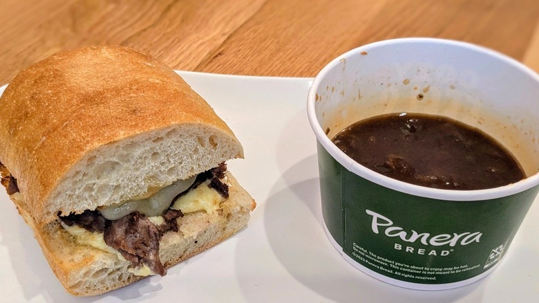 Half a steak sandwich next to a cup of French onion soup on a white plate