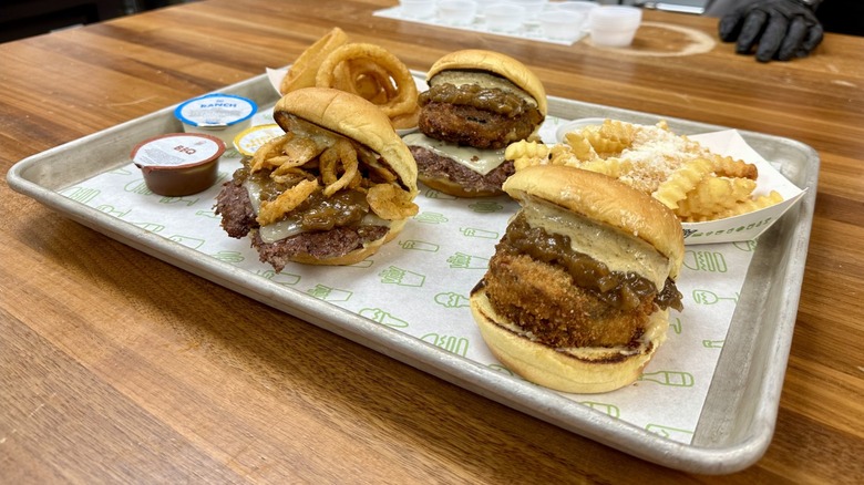 Shake Shack's French Onion Soup Burger, French Onion Soup 'Shroom Burger, French Onion Shack Stack, Garlic Parmesan Fries, Onion Rings, and dipping sauces on a metal tray with a placemat.