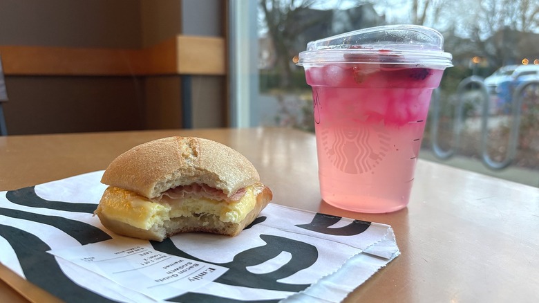 Starbucks Pink Cannon Ball drink next to bacon egg gouda sandwich on table in cafe