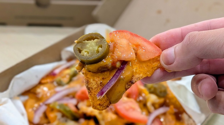 A hand holding a Dorito with cheese, onion, tomato, jalapeno, and hot honey