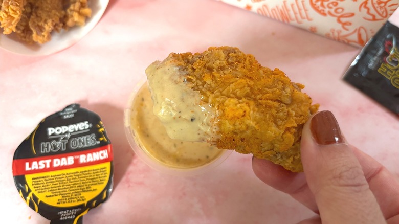 Hand holding Popeyes wing dipped in Last Dab Ranch sauce over pink surface