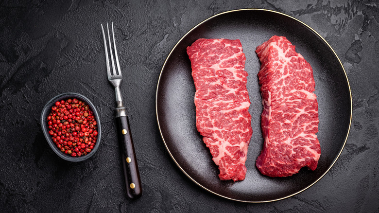 Two Denver cut beef steaks on black plate next to bowl of pink peppercorns on three pronged fork on black surface