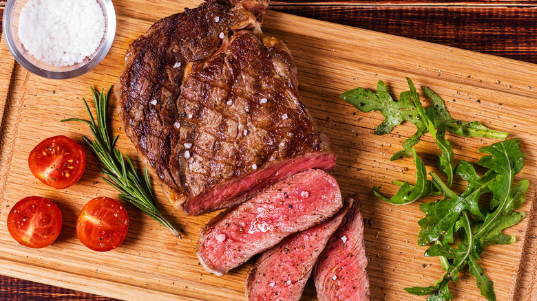 Grilled ribeye steak with crosshatch marks on wooden board next to tomatoes, arugula, rosemary, and salt