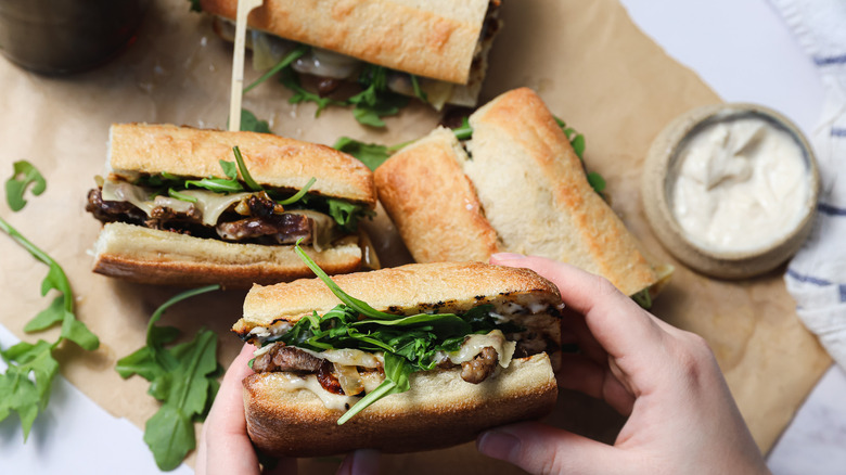 Hands holding a ribeye steak sandwich over more sandwiches on parchment