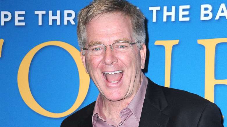 Rick Steves smiling in lavender shirt with black blazer and glasses