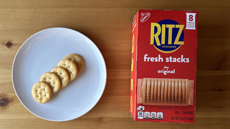 Five Ritz crackers on a white plate sitting on a wooden table top next to a red box of Ritz crackers