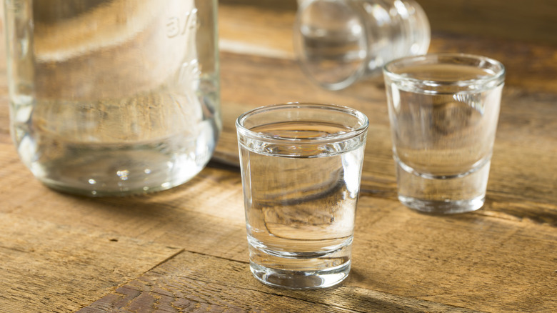 A bottle of clear alcohol resting on a wooden surface next to two full shot glasses of clear liquid