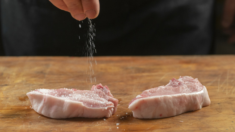 A hand sprinkles salt onto raw meat on a counter-top for a dry brine