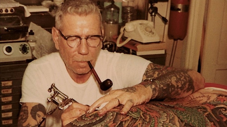 Old photo of Norman Collins tattooing someone while smoking a pipe