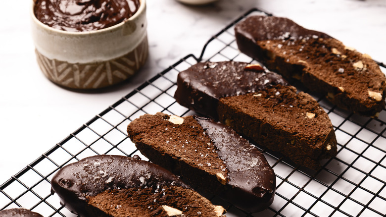 Four chocolate biscotti on a rack with ganache in background.