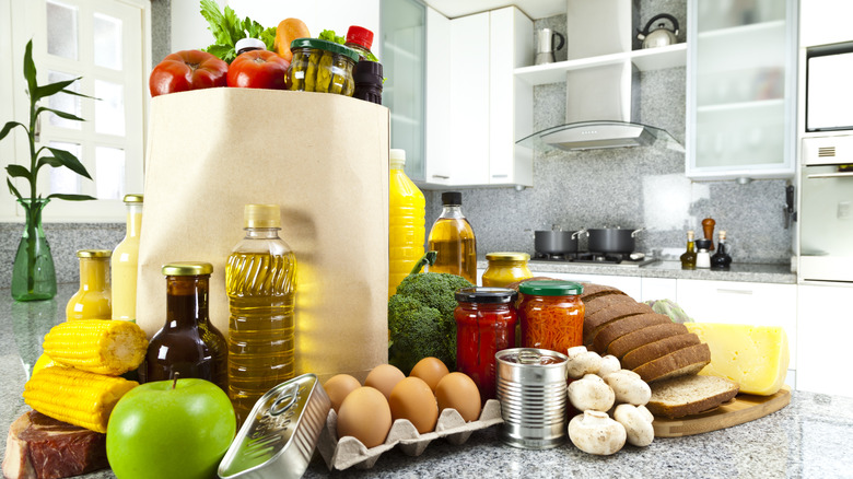 A kitchen countertop full of groceries (fruit, vegetables, bread, eggs, cans, pasta sauce) spread about and also in brown paper bag