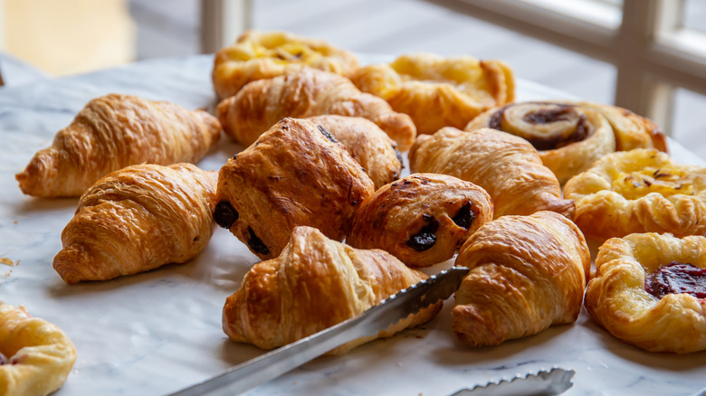 Breakfast pastries including croissants, pain au chocolat, and Danishes on wax paper next to silver tongs