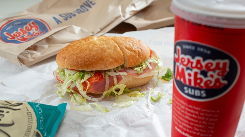 A Jersey Mike's sub on a paper wrapper beside a soft drink cup