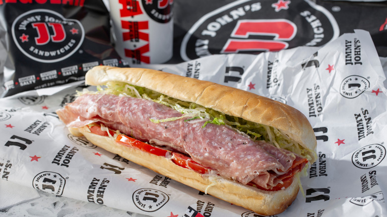 A Jimmy John's sub on a paper wrapper with other Jimmy John's packaging in the background