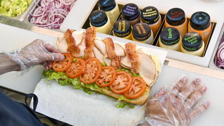 Gloved hands building a Subway sandwich on a white counter with condiments