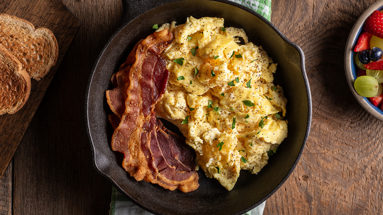 scrambled eggs in cast iron pan with bacon and side of toast and fruit